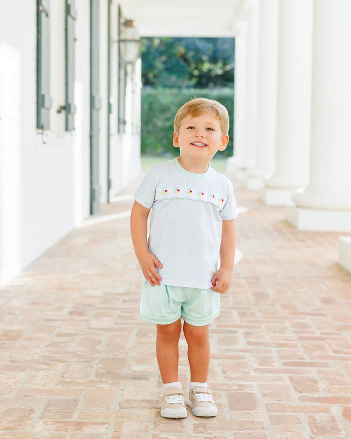 A young boy stands on a brick walkway in front of a white building with columns, wearing a white short-sleeve t-shirt with a small red embroidered pattern across the chest and light mint green shorts, that store