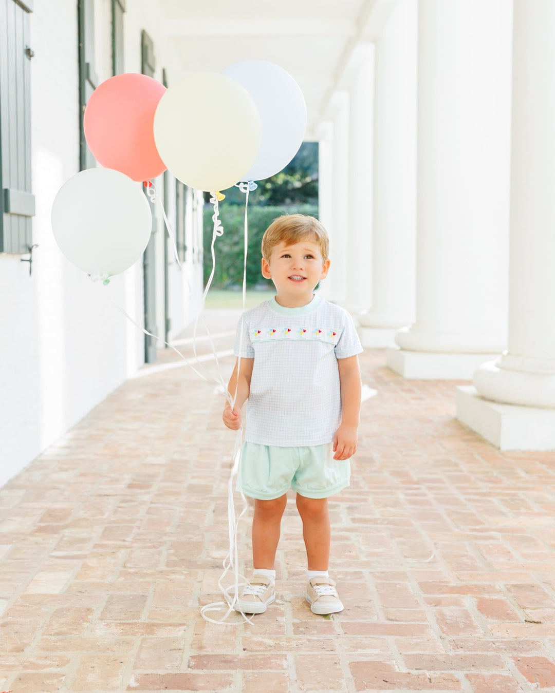 A young toddler boy stands on a brick porch with white columns, holding a cluster of pastel balloons. He is wearing a white short-sleeve shirt with a small embroidered pattern across the chest and light mint green shorts, that store