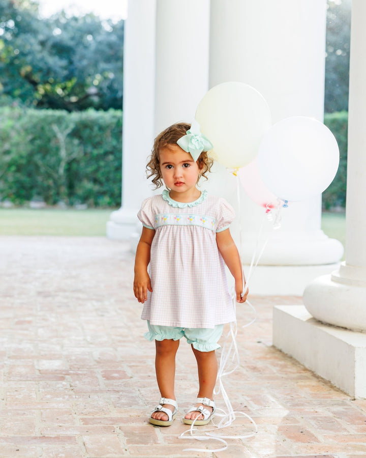 A toddler girl stands on a stone walkway in front of white columns, wearing a pale pink smocked dress with a mint green ruffled hem, a mint headband, and holding large white balloons, that store