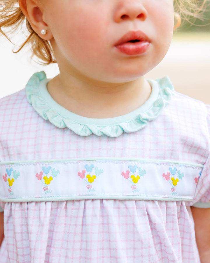 Close-up of a child wearing a light pink and white gingham dress with a mint green ruffled collar and a decorative embroidered band across the chest, that store