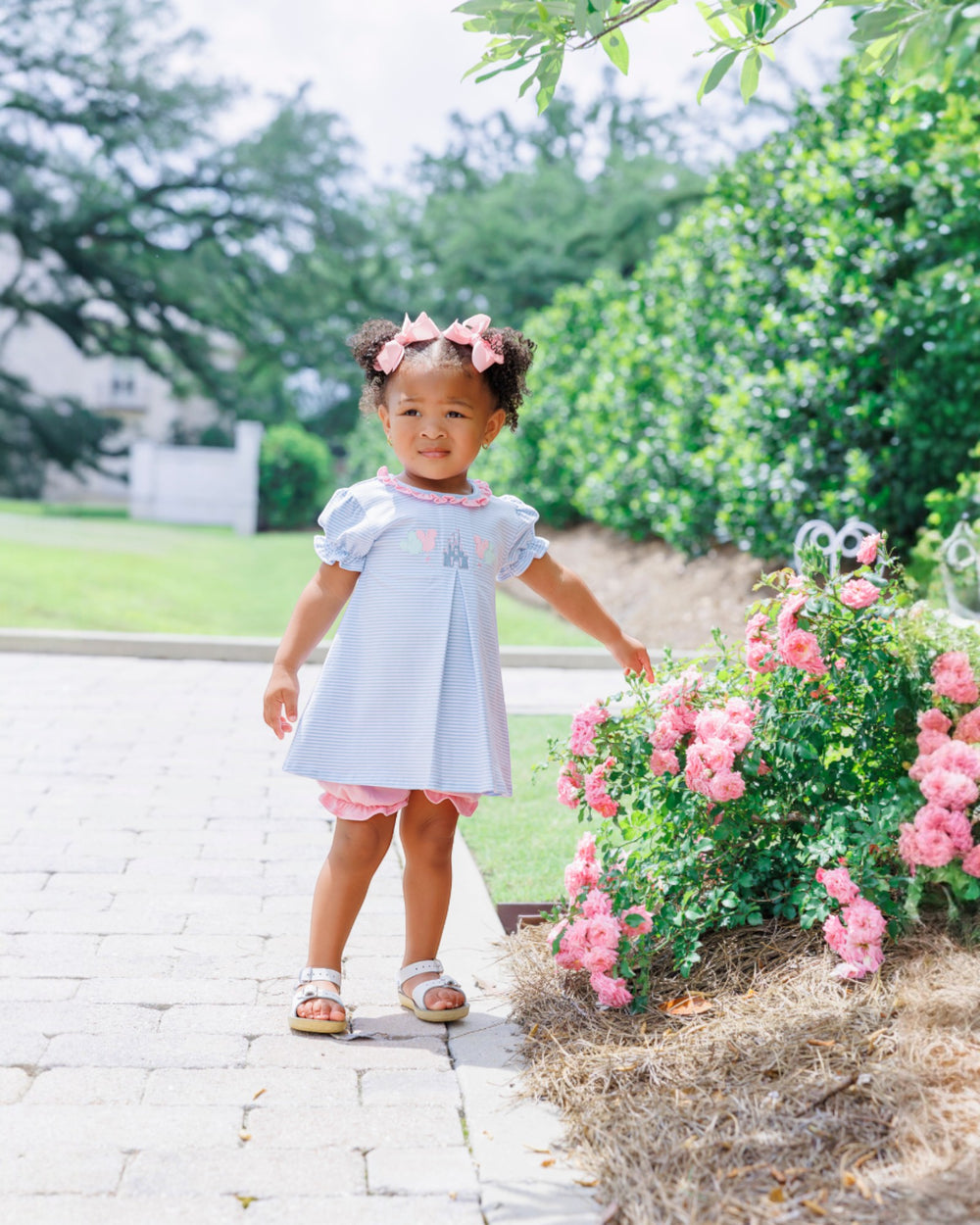 Young girl in a light blue dress standing on a stone path with pink flowers and greenery around her.
