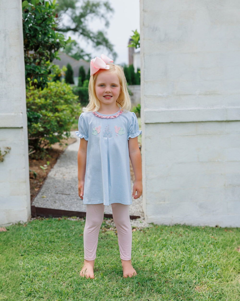 Young girl in a light blue dress and pink pants standing outdoors near a stone wall.