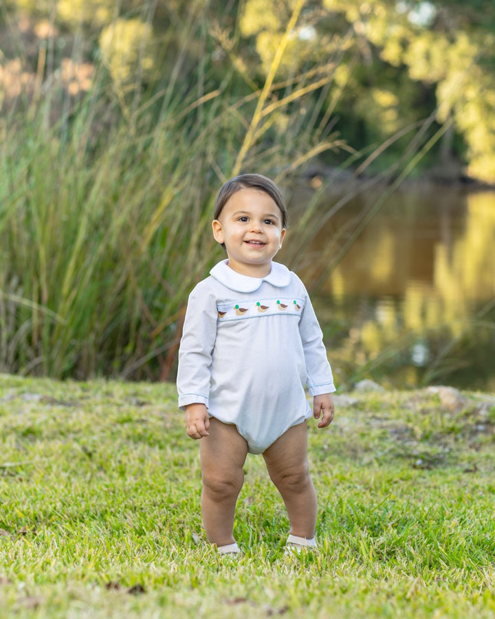 A toddler standing outdoors on grass near water, wearing a light blue long-sleeve baby bodysuit with a white collar and small embroidered details across the chest, that store