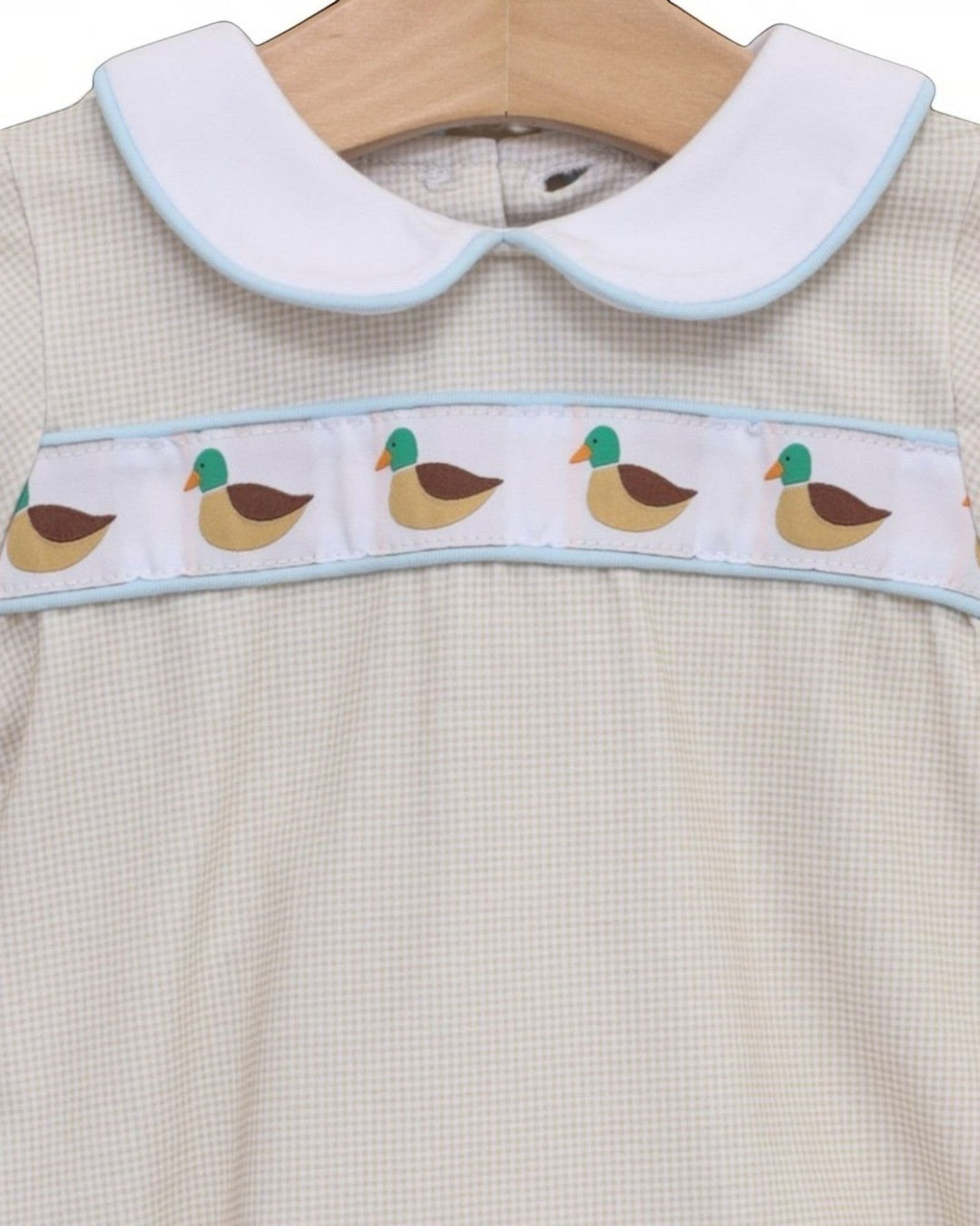 A close-up of a baby dress featuring a white Peter Pan collar with light blue trim, a white chest band decorated with a repeating duck pattern, and a cream-colored gingham fabric body, that store
