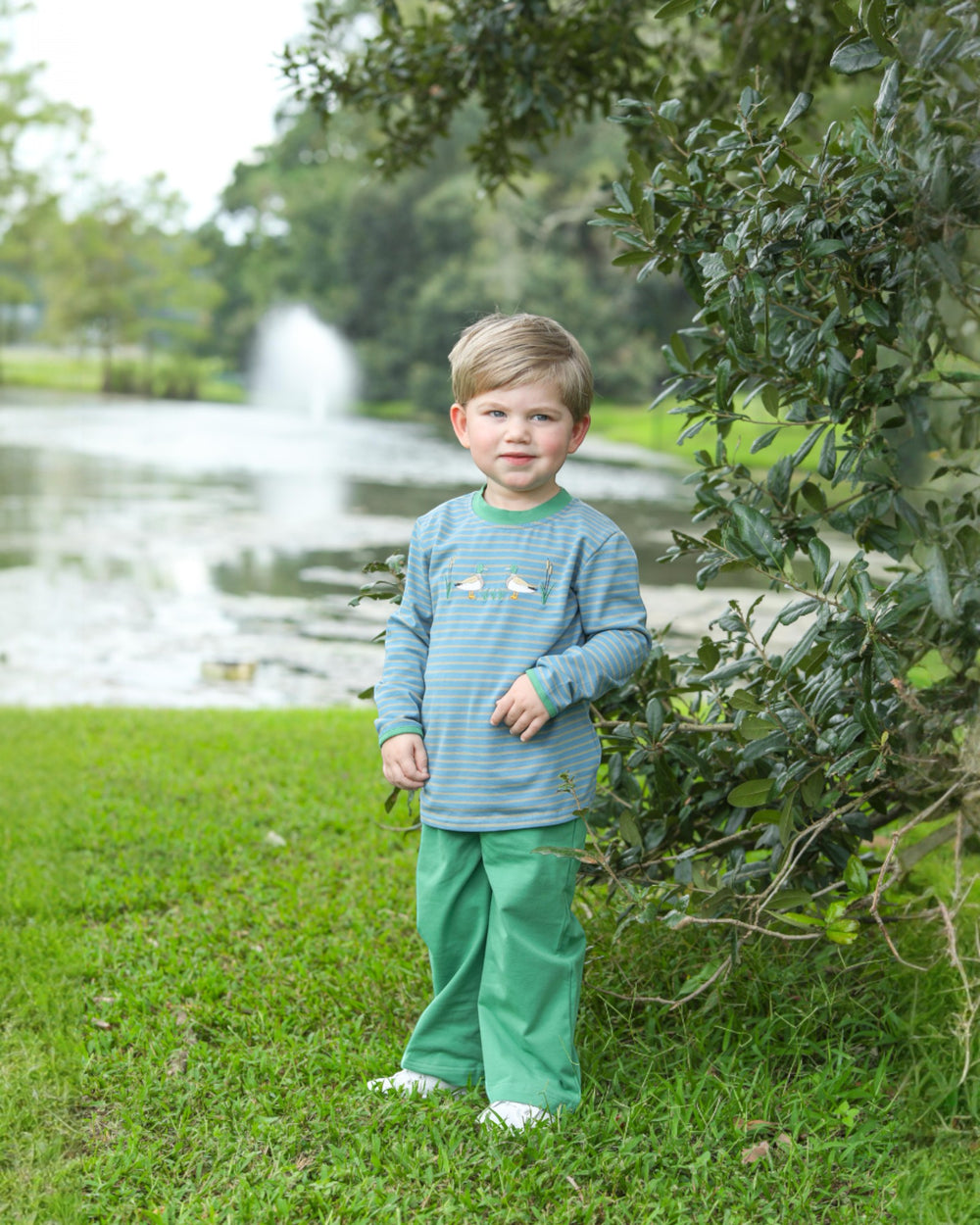 Child in a green outfit standing outdoors with a pond and trees in the background