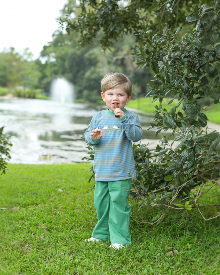 Child in a blue shirt and green pants standing outdoors with a lake and trees in the background