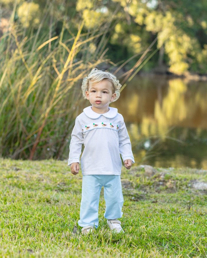 A toddler standing outdoors on grass near a pond, wearing a white long-sleeve top with a Peter Pan collar and small colorful embroidered details across the chest, paired with light blue pants and white shoes, that store
