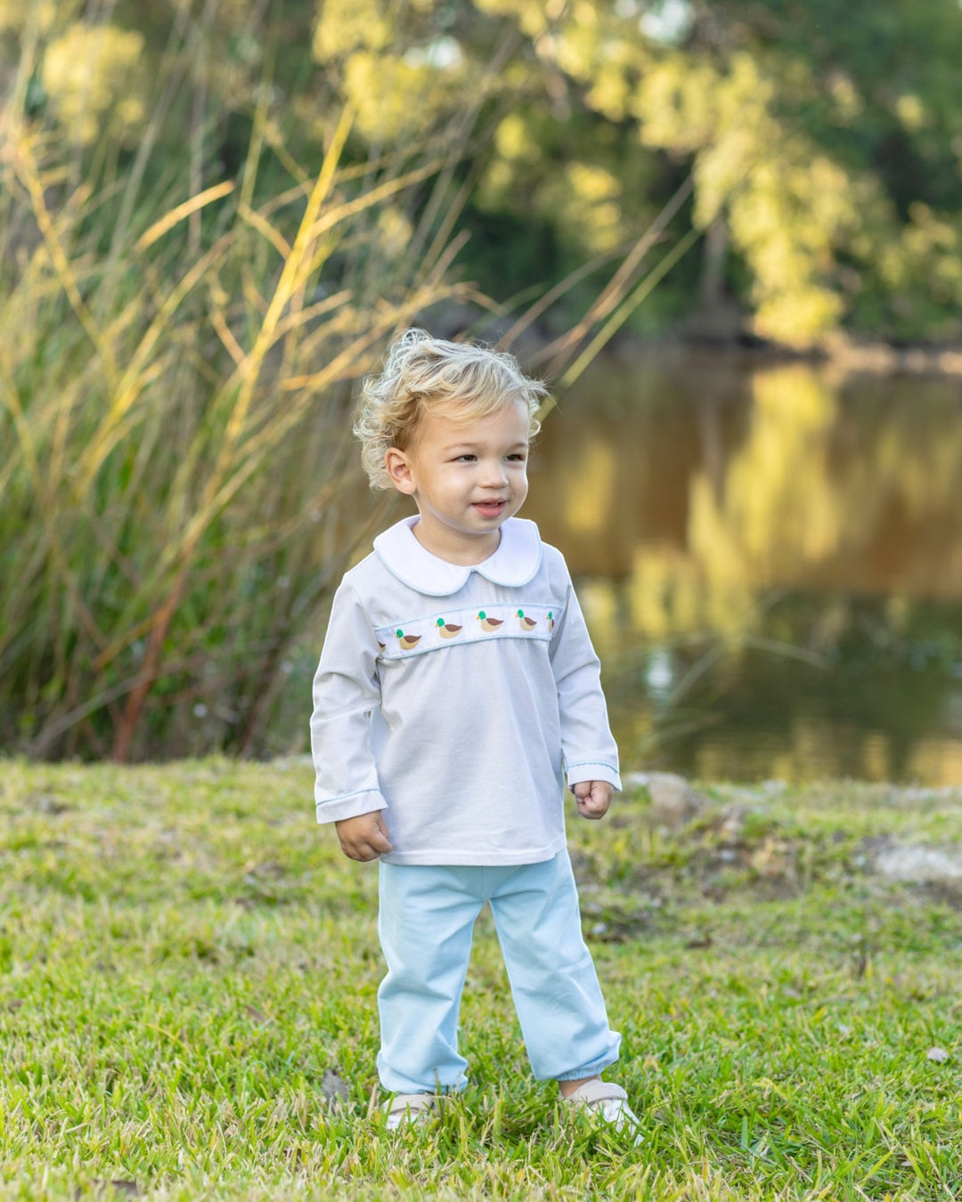A toddler standing outdoors on grass near a pond, wearing a white long-sleeve top with a Peter Pan collar and small embroidered details across the chest, paired with light blue pants, that store