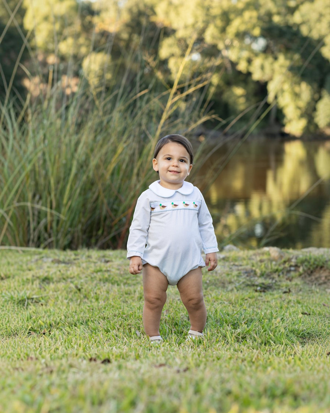 A toddler standing on grass outdoors near a pond, wearing a light blue long-sleeve romper with small embroidered details across the chest, that store