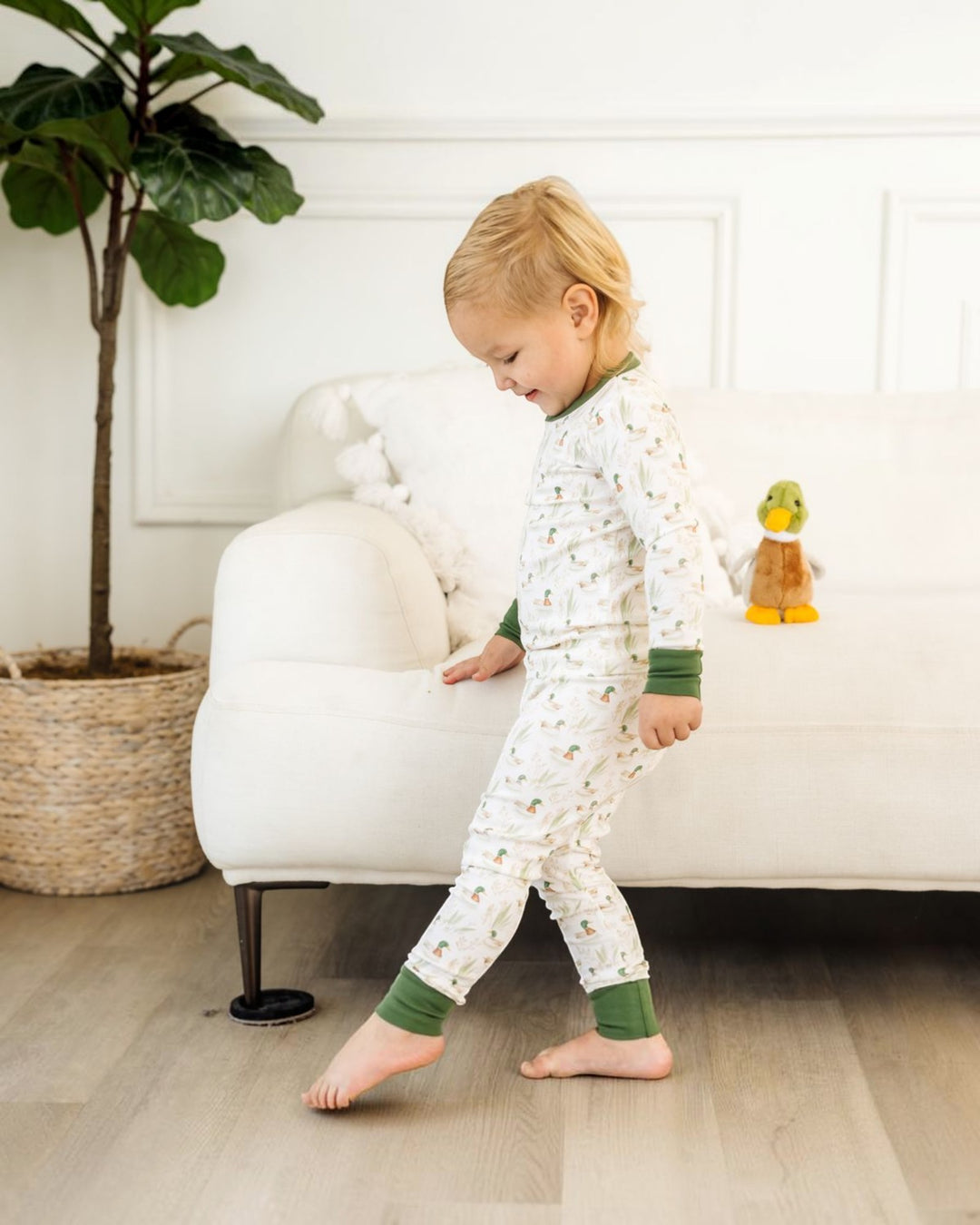 A toddler walking barefoot in a bright room, wearing a white long-sleeve pajama set with a small repeating pattern and green cuffs and collar, that store
