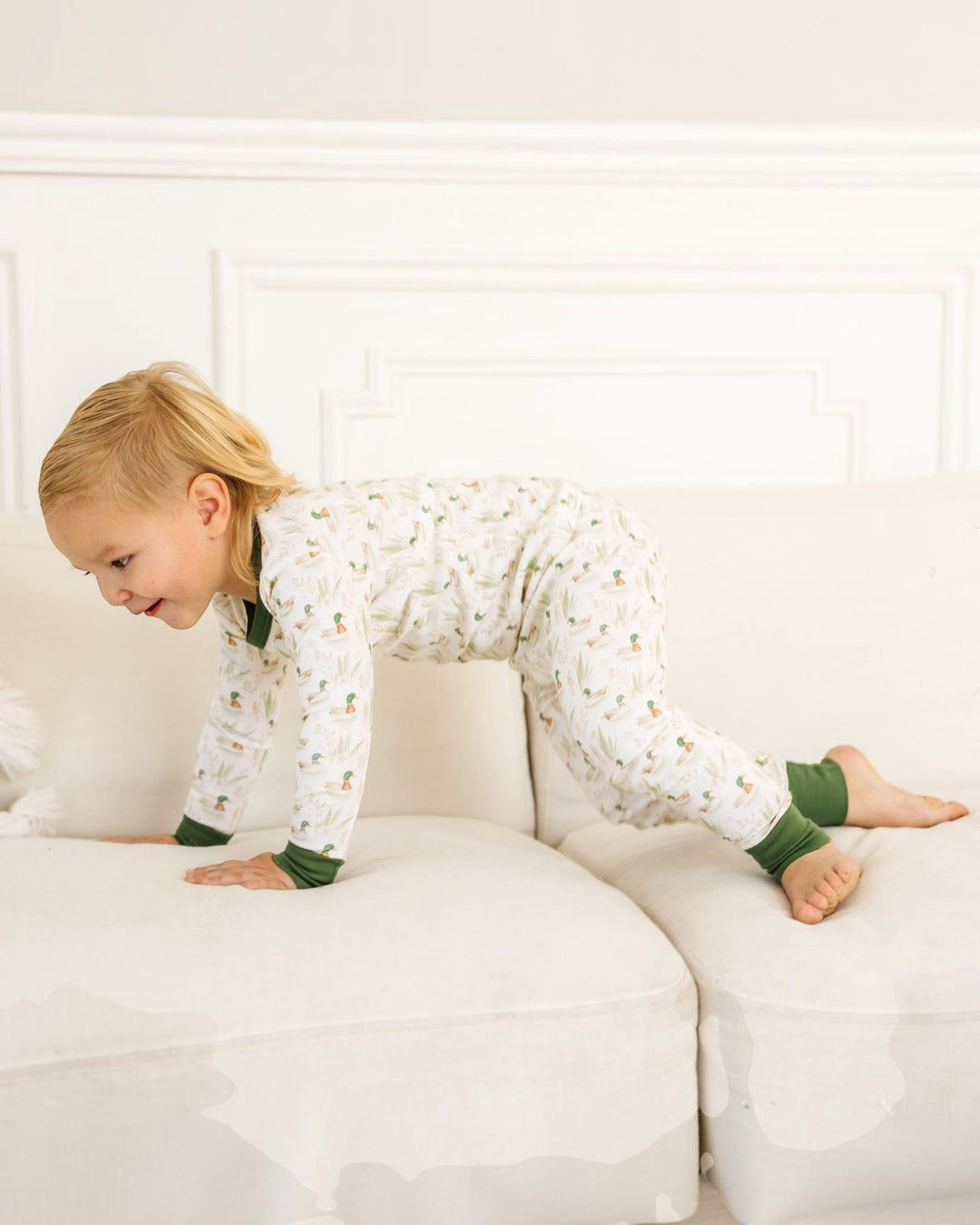 A young child wearing a white long-sleeve pajama set with a small green and orange pattern and solid green cuffs and collar, crawling on a white sofa in a bright room, that store
