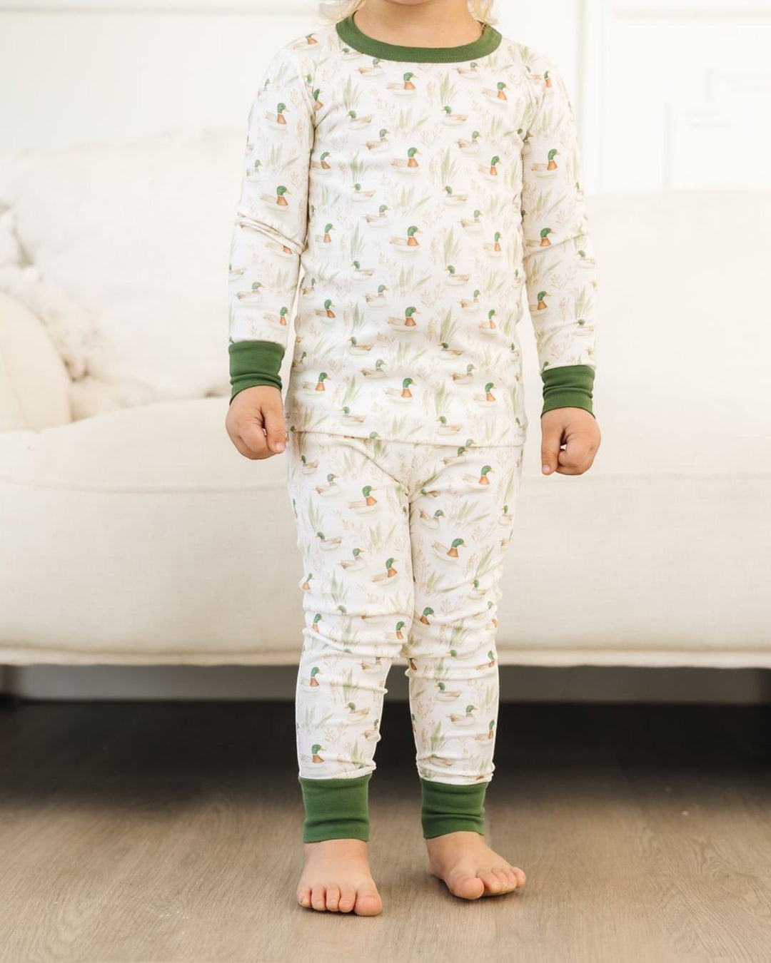 A child wearing a long-sleeve pajama set with a white background, green cactus and pinecone print, and solid green ribbed collar, cuffs, and ankle cuffs, standing barefoot on a wood floor in front of a white sofa, that store