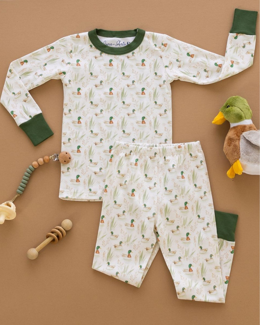 A baby long-sleeve pajama set laid flat on a tan background, featuring an off-white fabric with a green and orange cactus print and solid green collar, cuffs, and knee patches. The set includes matching pants, that store