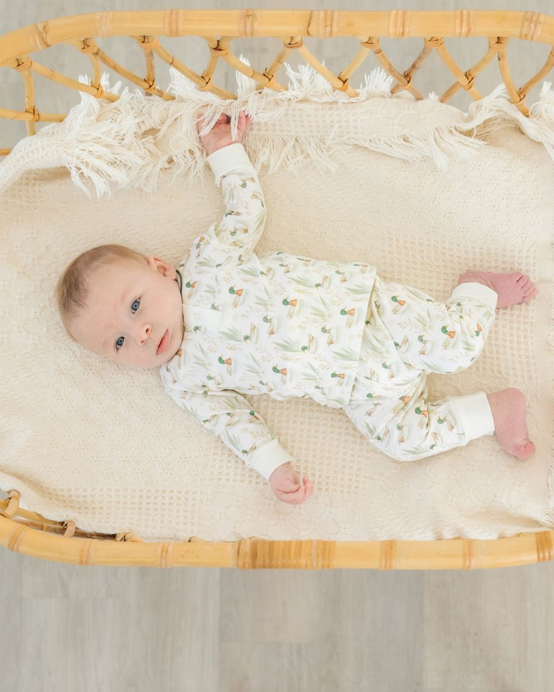 A baby lying on their back in a woven rattan bassinet, wearing a white long-sleeve pajama set with a delicate green and orange botanical pattern. The bassinet is lined with a cream-colored textured blanket with fringe trim, that store