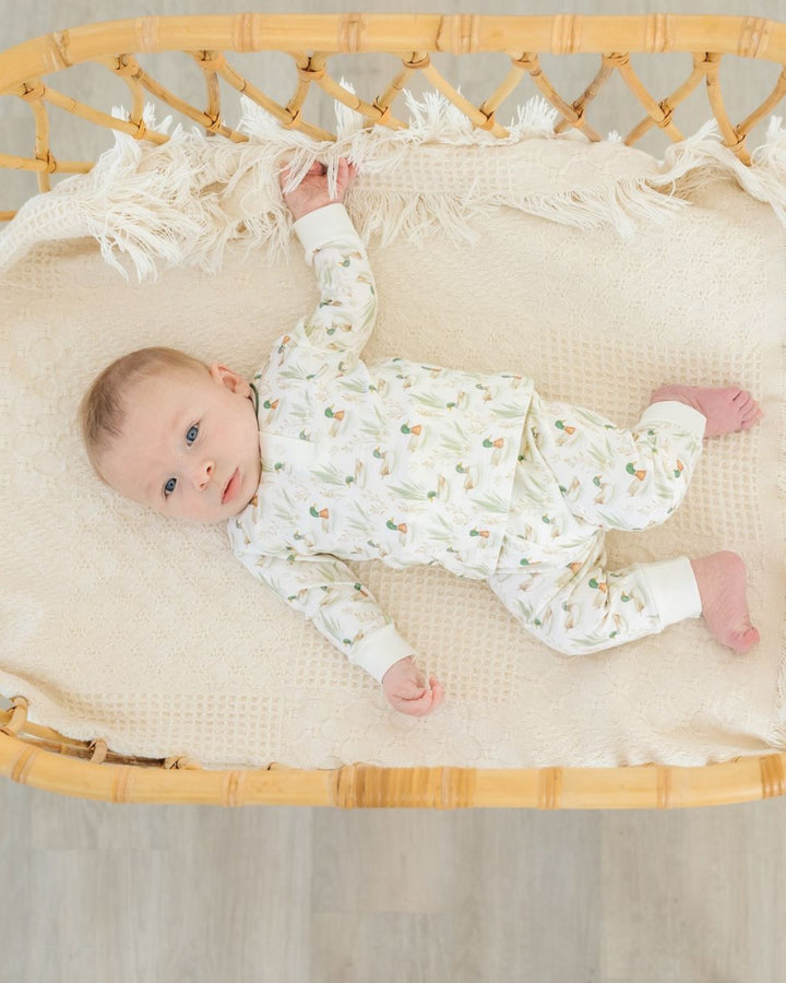 A baby lying on their back in a woven rattan bassinet, wearing a white long-sleeve pajama set with a delicate green and orange botanical pattern. The bassinet is lined with a cream-colored textured blanket with fringe trim, that store