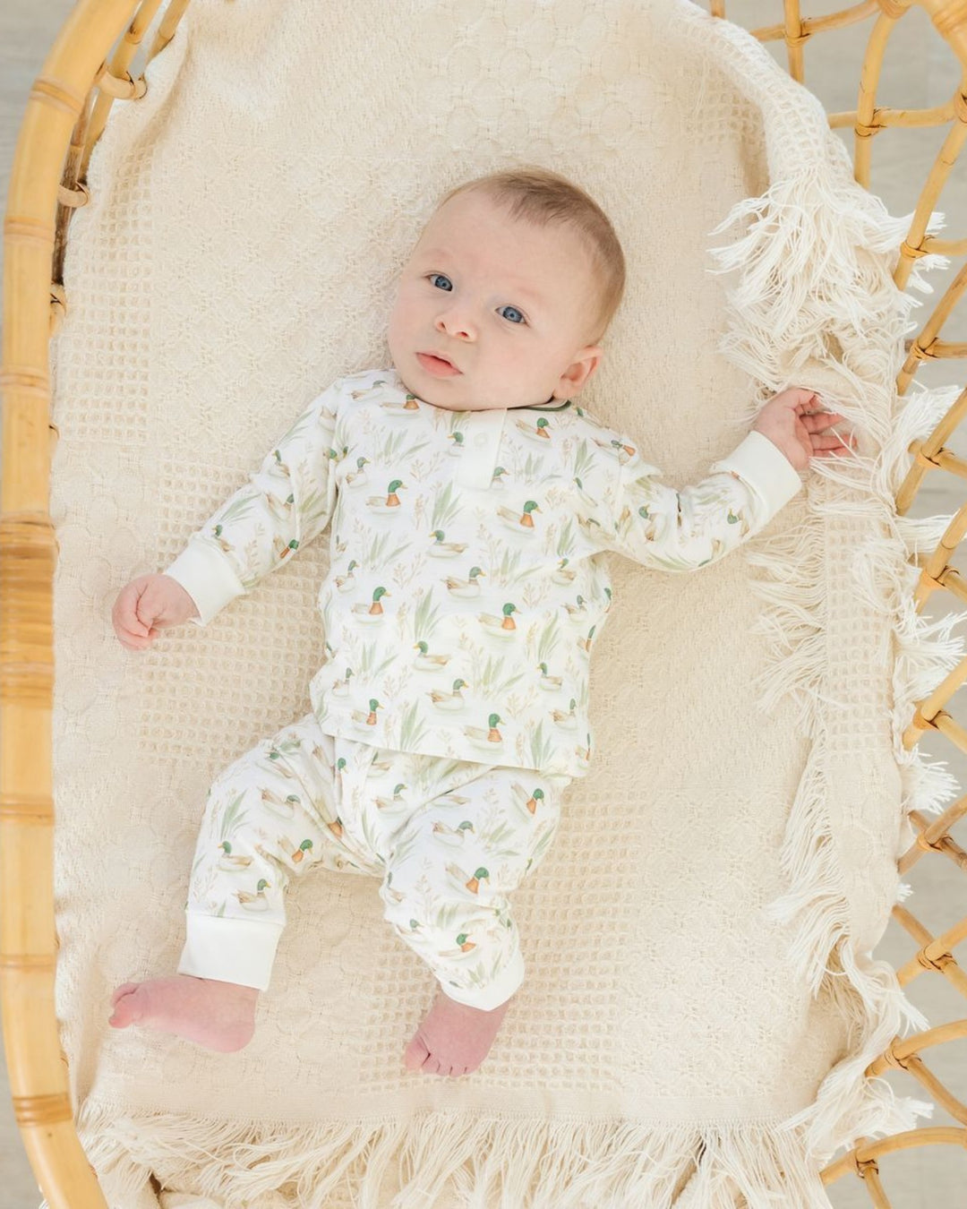 A baby lying on a cream woven bassinet, wearing a matching long-sleeve pajama set with a green palm tree and orange pineapple print on a white background, that store