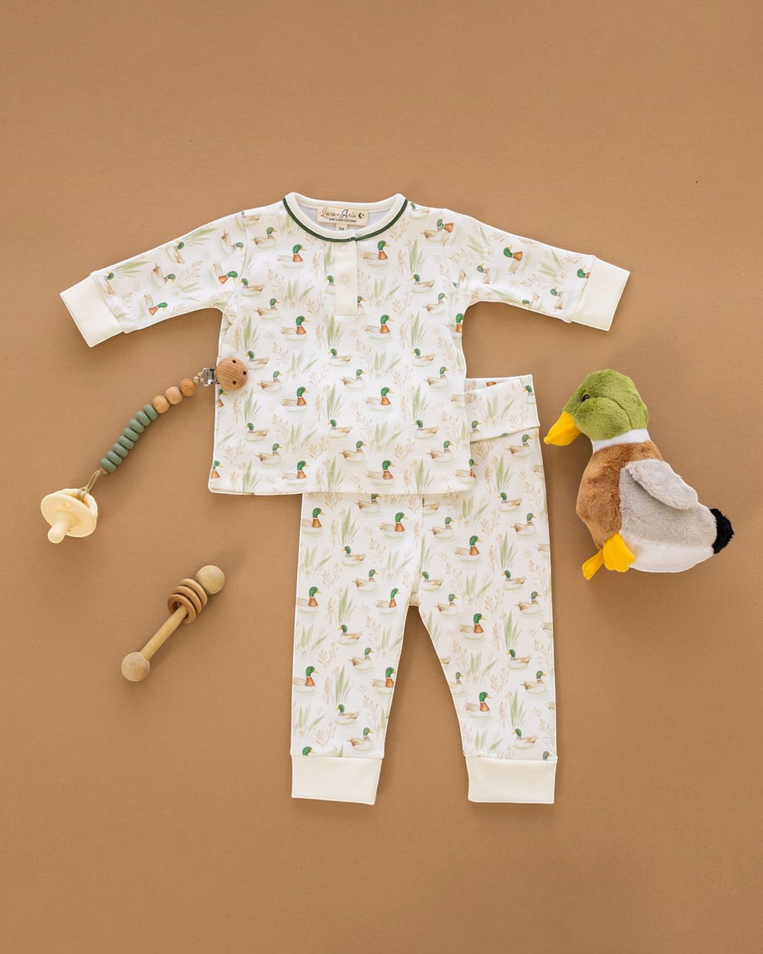 A baby pajama set laid flat on a tan background, including a long-sleeve top and matching pants in white with a small green and orange pineapple print. A plush duck toy and wooden baby teething toys are placed beside the pajamas, that store