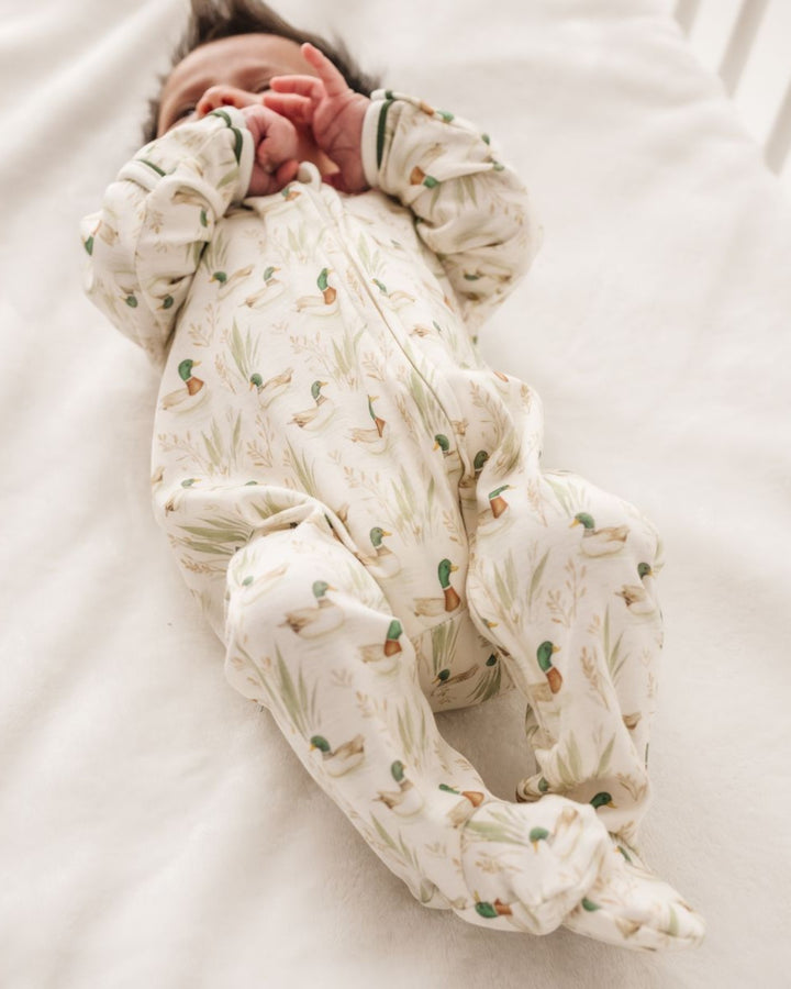 A newborn baby lying on a white blanket, wearing a long-sleeve footed sleeper with a duck and reeds pattern on an off-white background, that store