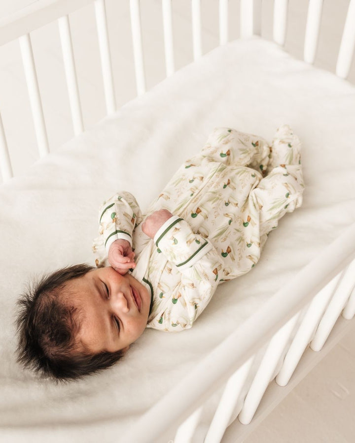 A newborn baby sleeping peacefully on their back in a white wooden crib, wearing a cream-colored footed sleeper with a green and orange botanical pattern, that store