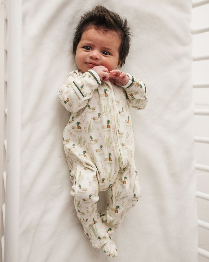 A newborn baby lying on their back on a white mattress, wearing a cream-colored footed sleeper with a green and orange cactus pattern, that store