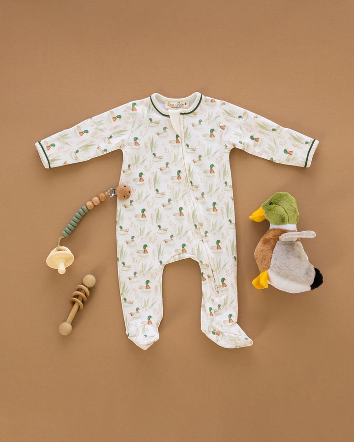 A baby footed sleep sack laid flat on a warm brown background, featuring a white base with a green and orange pineapple print and dark green trim. It is styled with baby toys, including a wooden teething toy, a pacifier clip, and a plush duck, that store