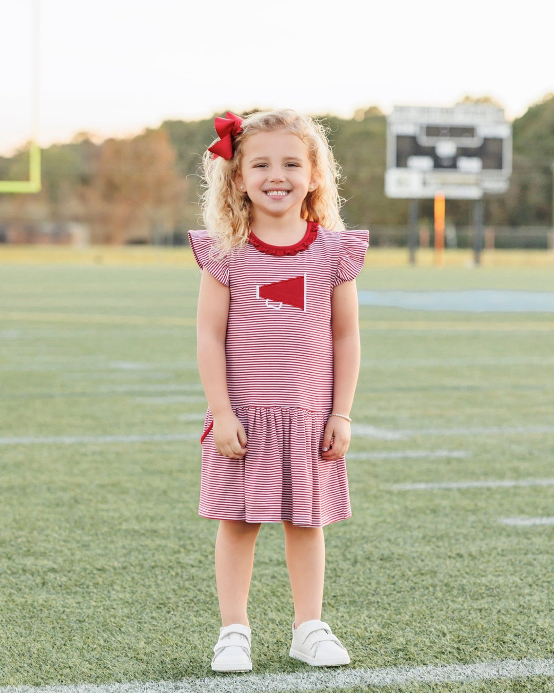 Young girl in a red and white dress with a red bow standing on a sports field.