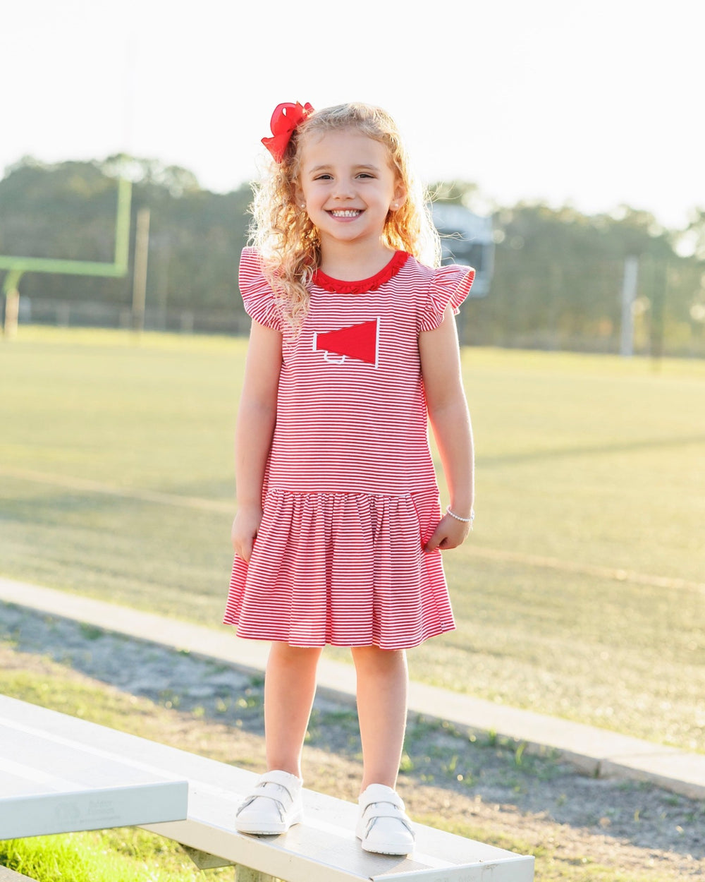 Young girl in a red and white dress standing on a wooden bench with a grassy field in the background.