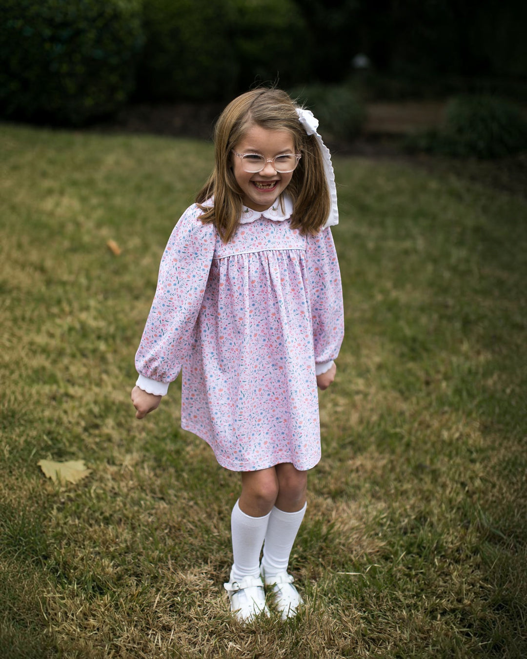 A young girl with glasses and a white hair bow stands on a grassy lawn wearing a light pink long-sleeve dress with a tiny blue and red floral print, white socks, and white shoes, that store