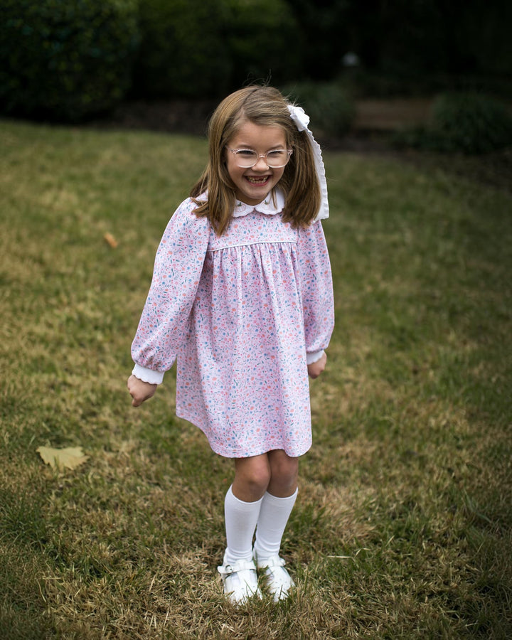 A young girl with glasses and a white hair bow stands on a grassy lawn wearing a light pink long-sleeve dress with a tiny blue and red floral print, white socks, and white shoes, that store