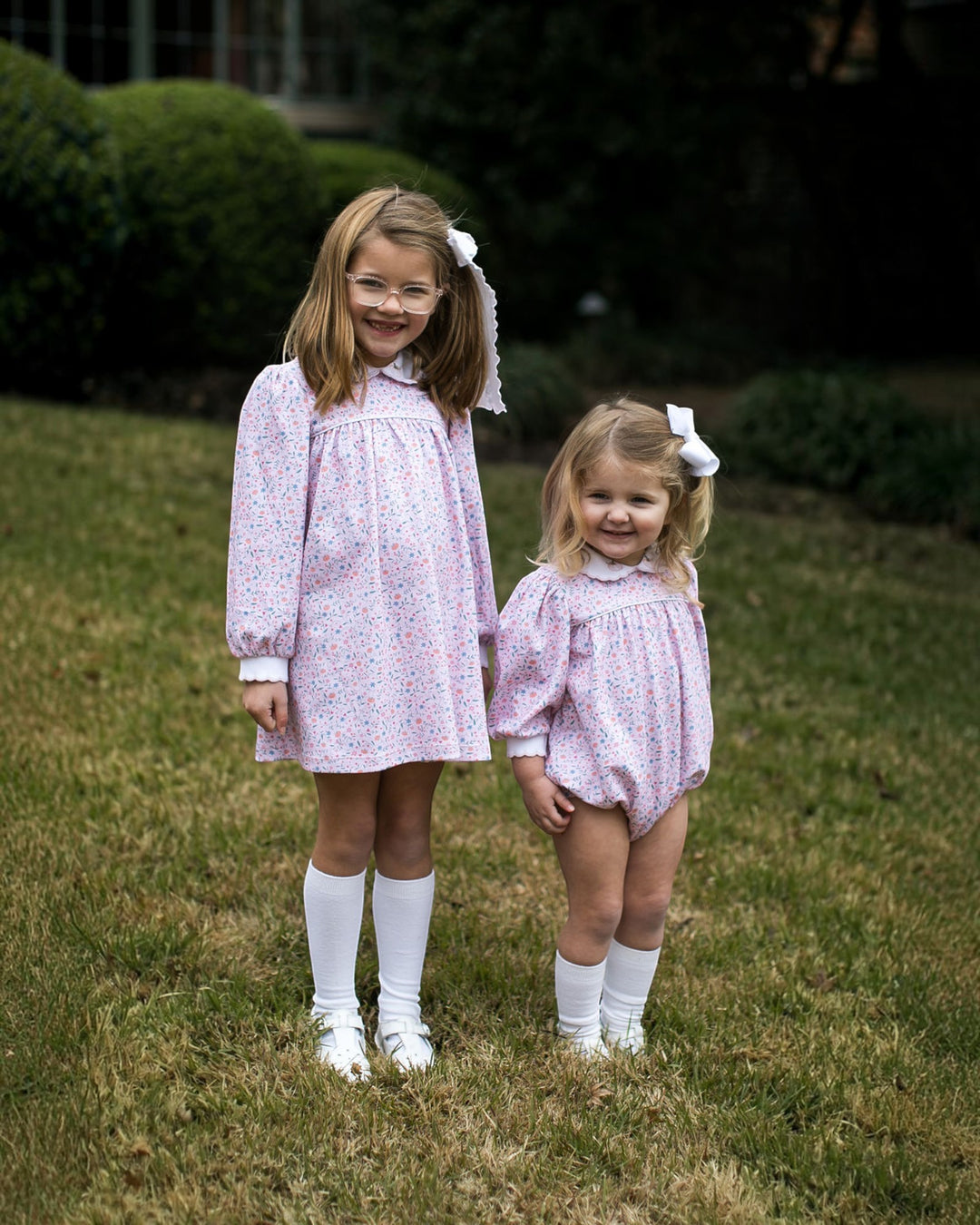Two young girls standing on a grassy lawn outdoors, wearing matching light pink floral babydoll dresses with white collars and cuffs, white knee-high socks, and white shoes. One girl wears a long dress and the other wears a short romper, both with white bows in their hair, that store