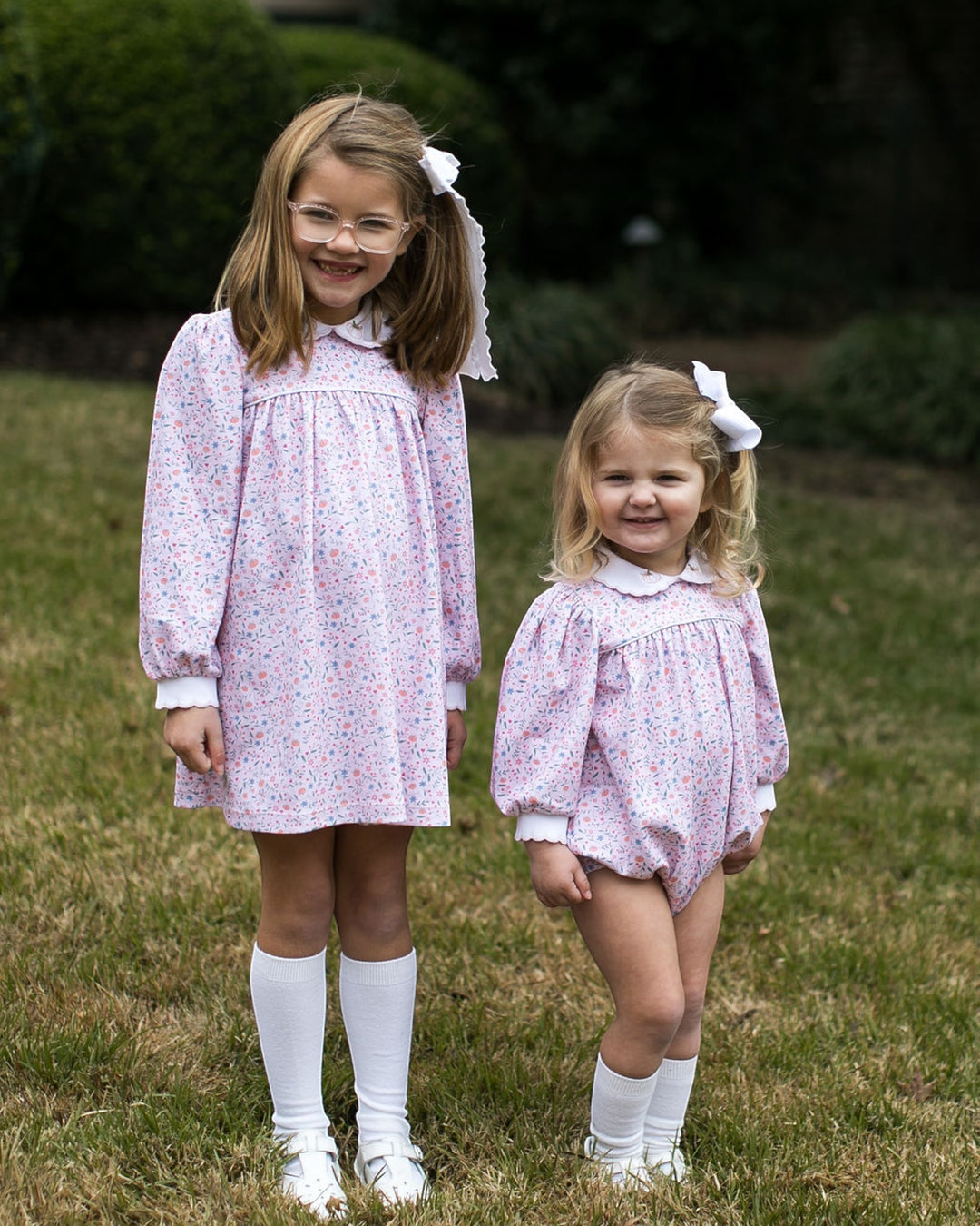 Two young girls standing outdoors on grass, wearing matching long-sleeve white dresses with a small pink and blue floral print, white collars, white knee-high socks, and white shoes. The girl on the left wears glasses and a white hair bow, and the girl on the right wears a white hair bow, that store