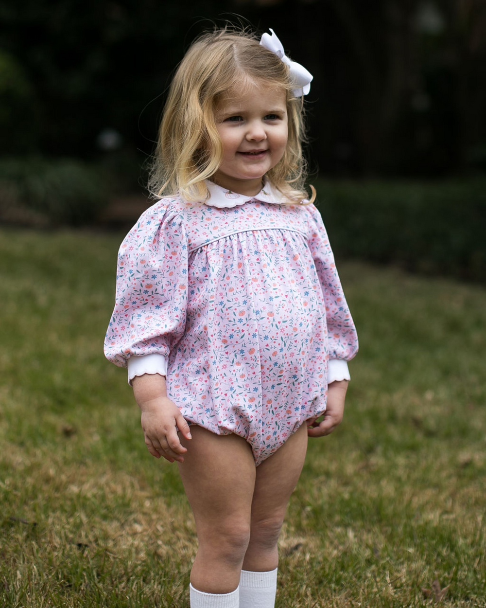 A young toddler girl with blonde hair and a white bow stands outdoors on a grassy lawn, wearing a long-sleeve floral bubble romper in pink, blue, and white, paired with white socks, that store
