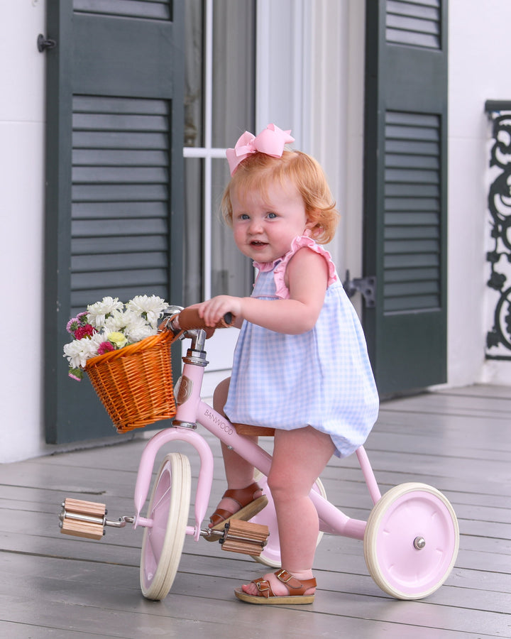 Child on a pink tricycle with a basket of flowers on a wooden deck.