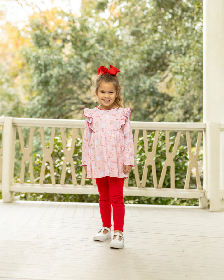 Young girl in a pink dress and red leggings standing on a porch with greenery in the background