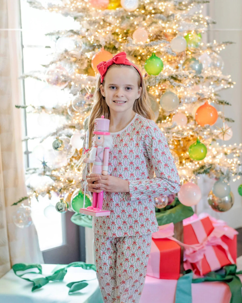 Young girl in pajamas holding a Nutcracker toy in front of a decorated Christmas tree with colorful ornaments and presents.