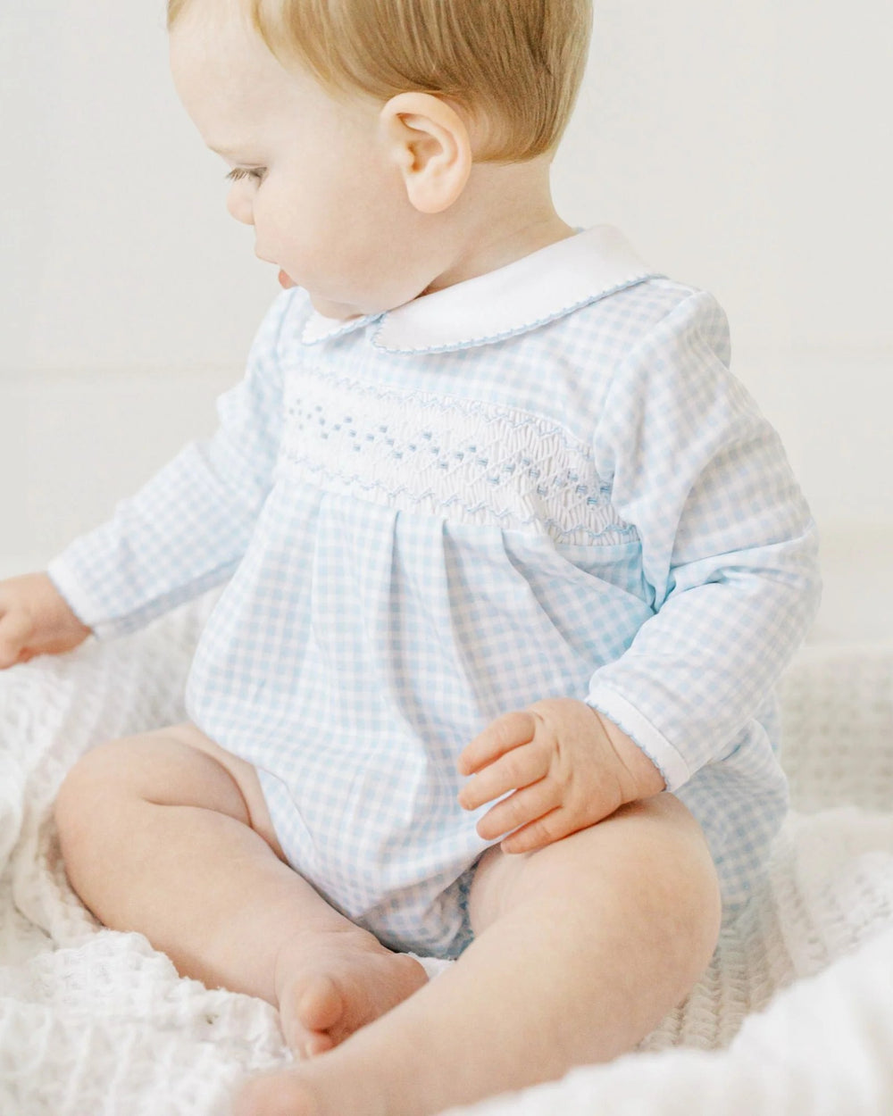 Baby wearing a light blue checkered outfit sitting on a white surface.