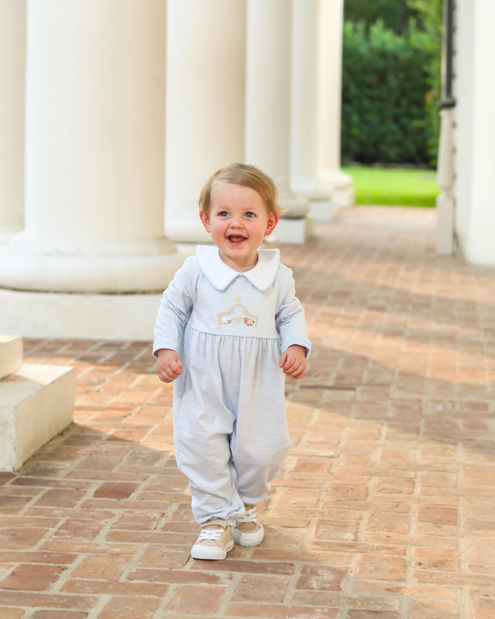 Baby in a light blue outfit standing on a brick path with columns in the background