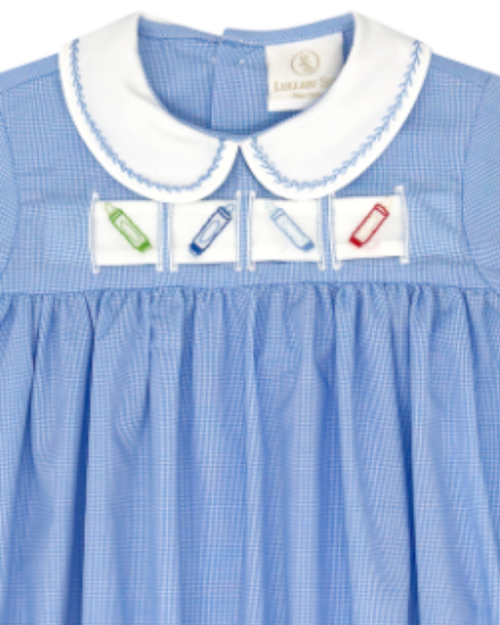 A close-up of a light blue gingham dress with a white Peter Pan collar and embroidered patches on the chest depicting colored pencils, that store