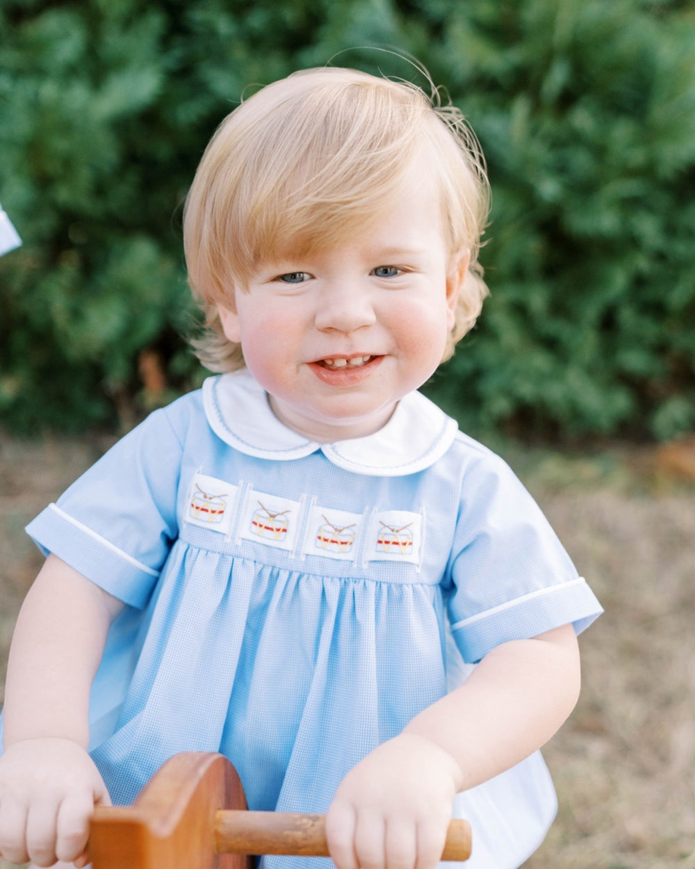 A young toddler with blonde hair wearing a light blue smocked dress with a white Peter Pan collar, smiling while holding a wooden toy outdoors, that store