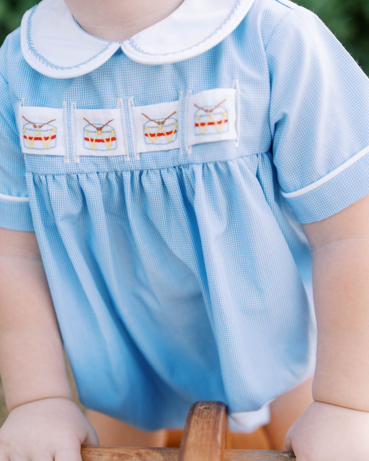 A close-up of a baby wearing a light blue gingham smocked dress with a white Peter Pan collar and embroidered drum motifs across the chest, that store