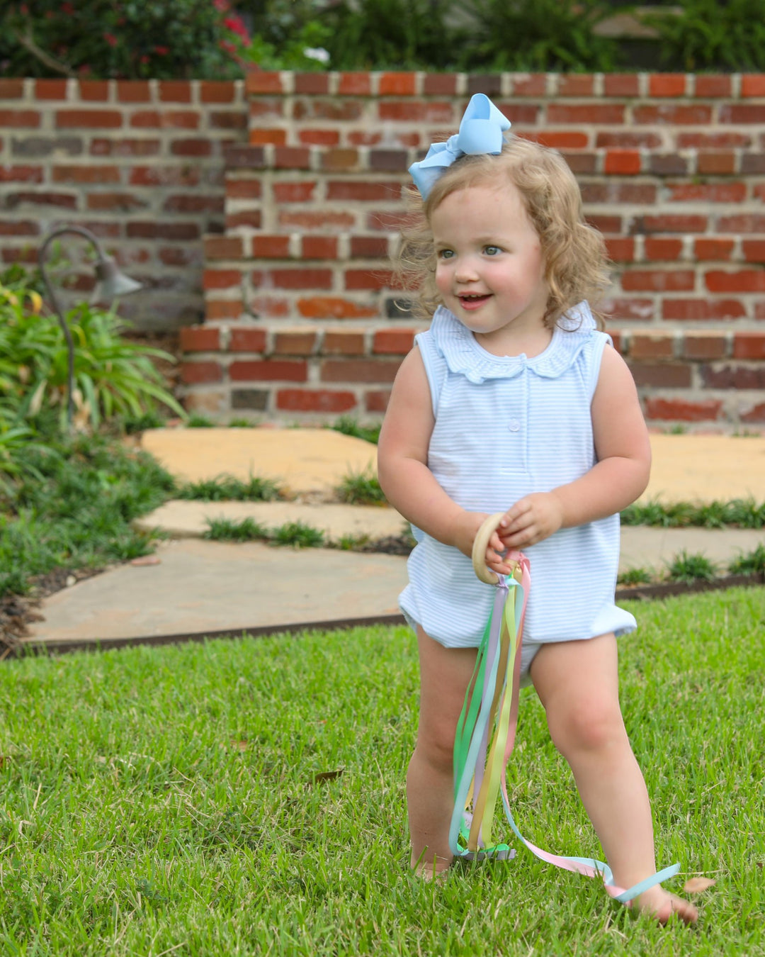 Young girl in a light blue outfit with a bow standing on grass in front of a brick wall.