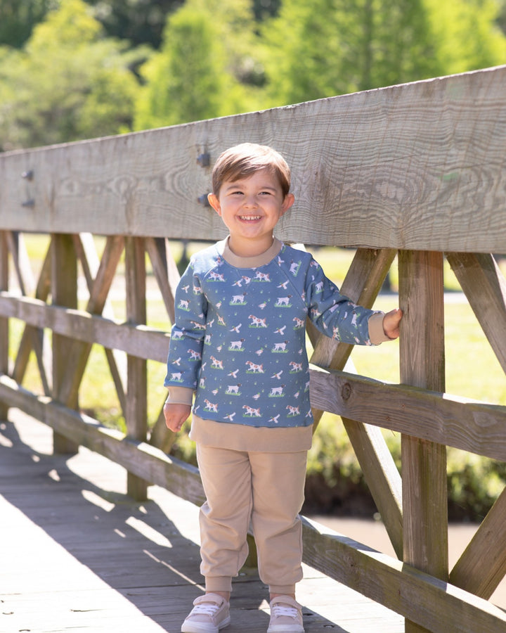 A young child standing on a wooden boardwalk bridge outdoors, wearing a blue long-sleeve shirt with a white animal print and beige pants, that store