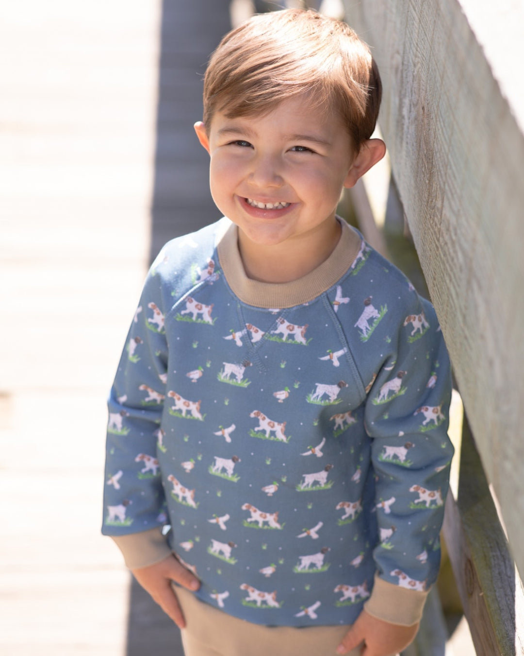 A smiling young boy wearing a blue long-sleeve crewneck sweatshirt with a cow print pattern, standing outdoors next to a wooden railing, that store
