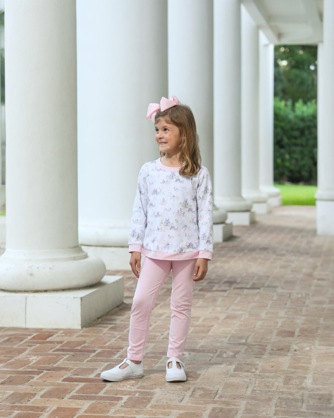 Young girl in a light-colored top and pink pants standing in front of white columns.