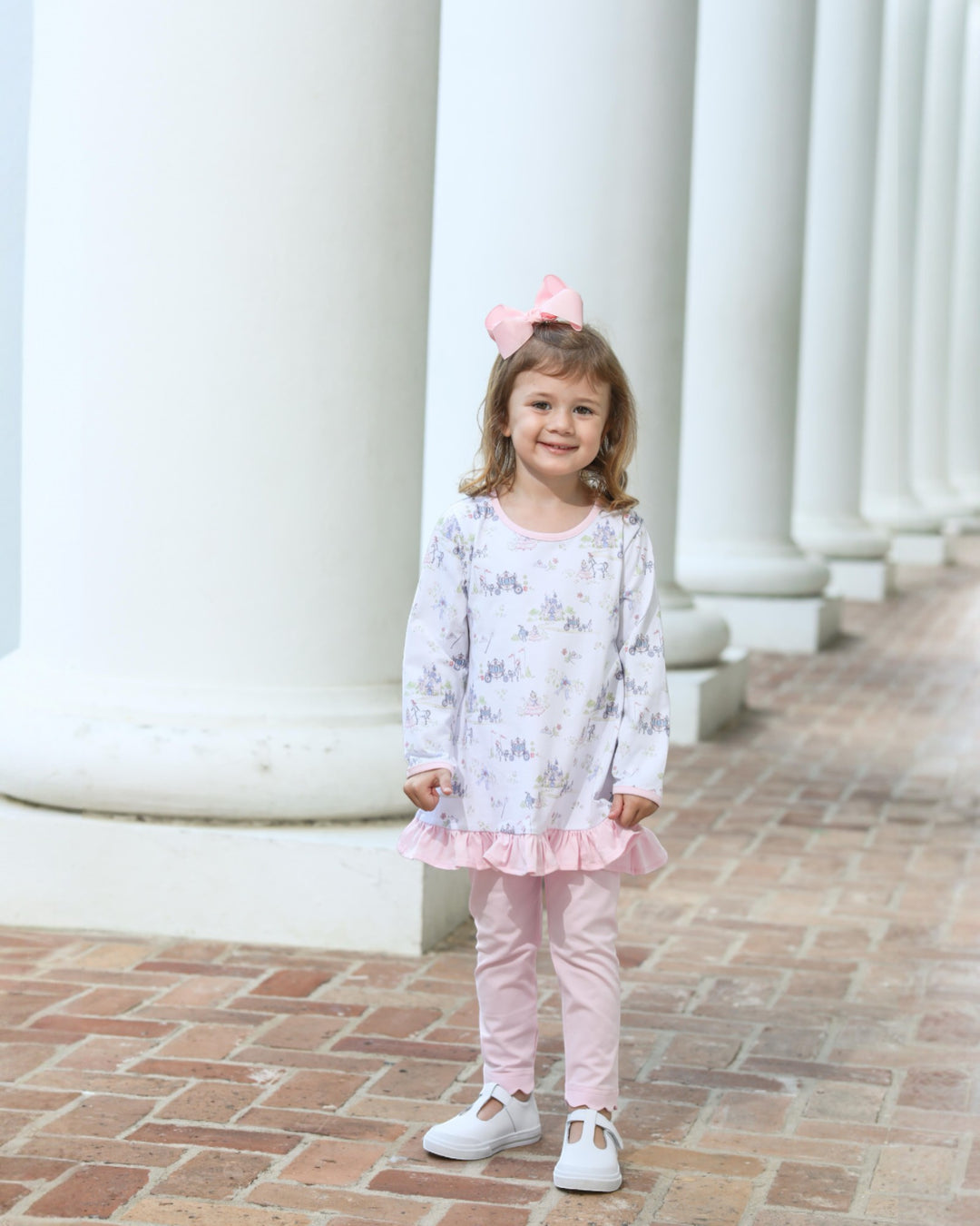 Young girl in a floral dress and pink pants standing in front of white columns.