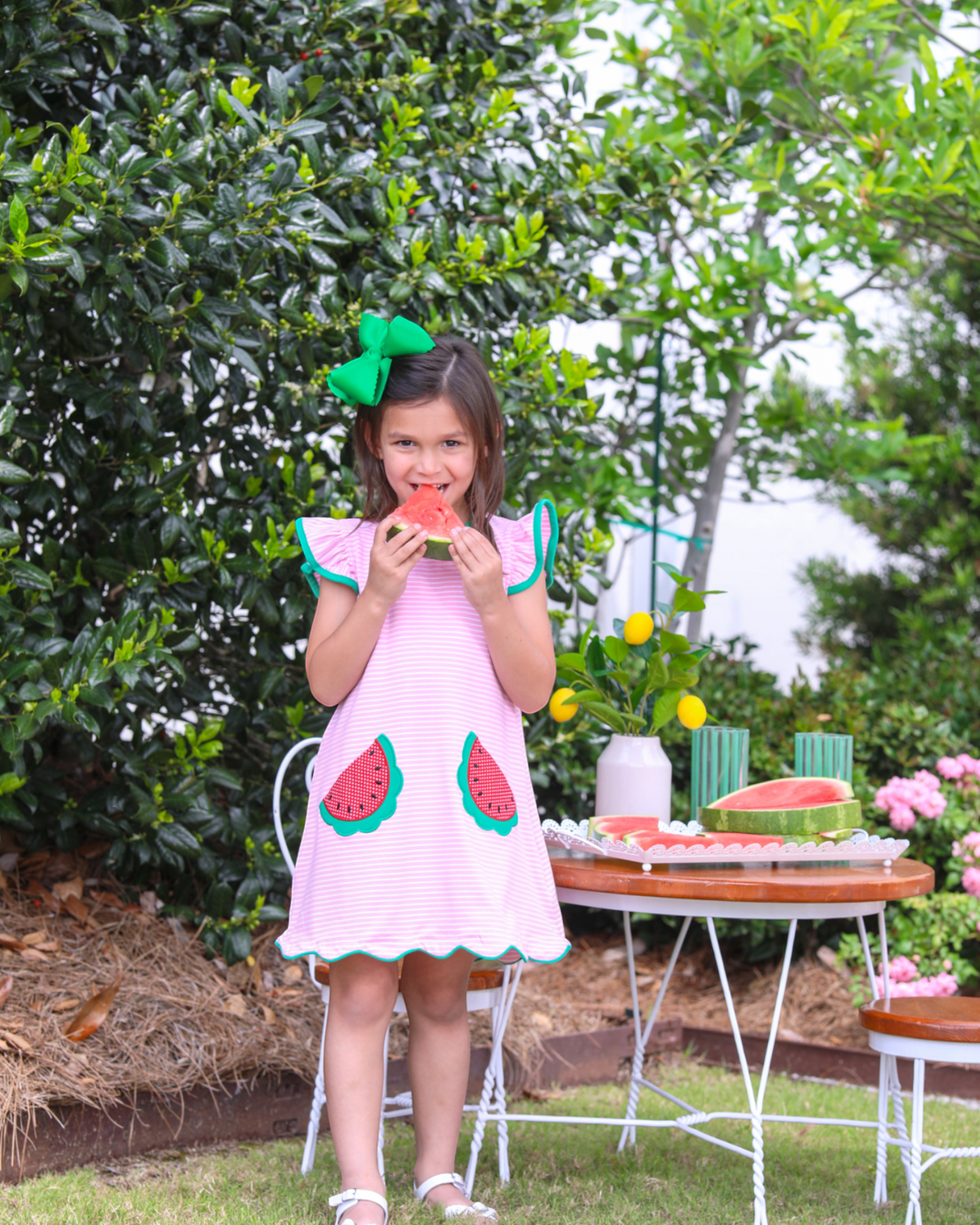Young girl in a pink dress with watermelon design eating watermelon outdoors.