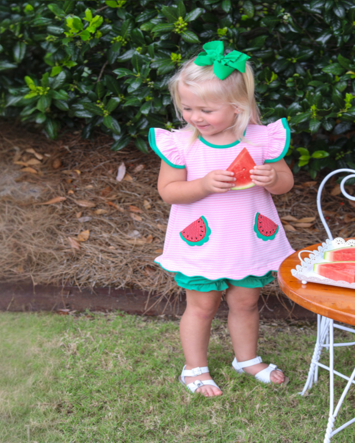 Young girl in a pink dress with watermelon design, holding a watermelon slice outdoors.