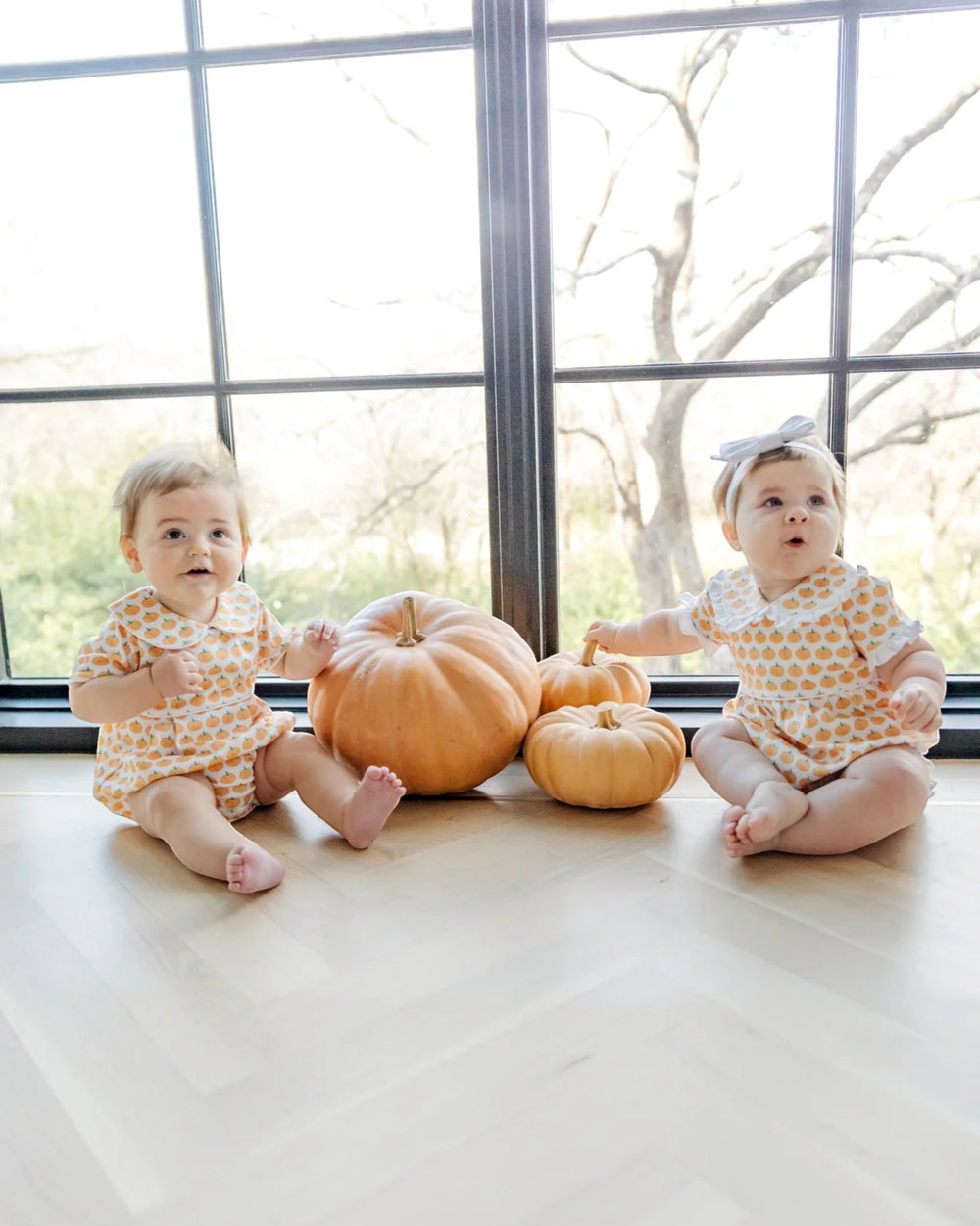Two babies in matching outfits sitting on a floor with pumpkins, in front of large windows.