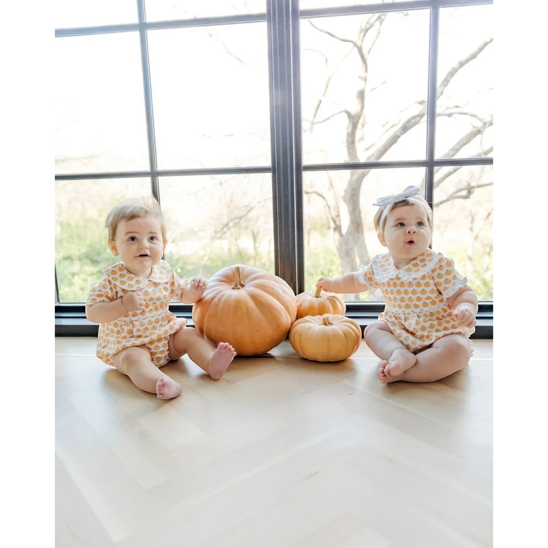 Two babies in matching outfits sitting on a floor with pumpkins, in front of large windows.