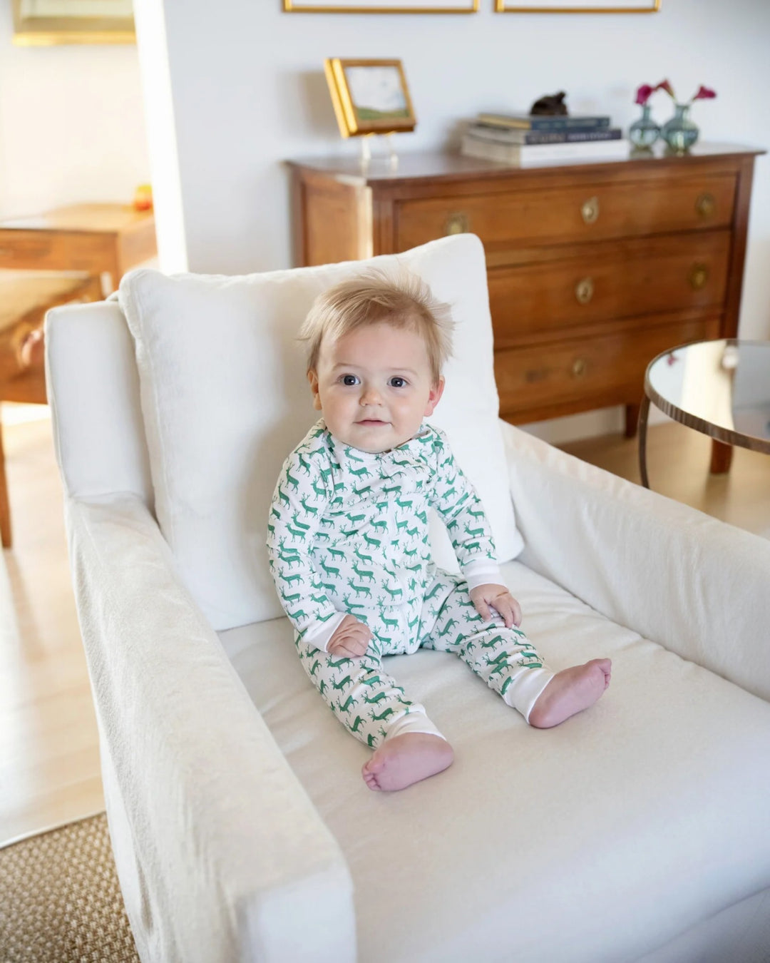 Baby in green and white pajamas sitting on a white chair in a room with wooden furniture.